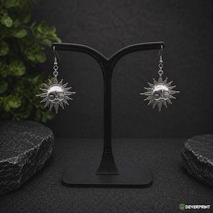 Puede incluir: Un par de pendientes colgantes de tono plateado con un diseño inspirado en el sol, que presenta un rostro central y rayos geométricos radiantes. Los pendientes cuelgan de ganchos plateados simples y se exhiben en un soporte negro.