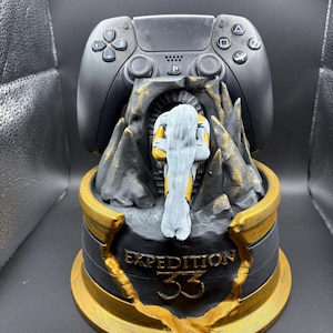 Peut inclure: Un support de manette de jeu noir avec une base sculptée représentant une silhouette en cape grise devant une arche sombre. La base est noire avec des accents dorés et le texte EXPEDITION 33 est gravé en lettres dorées sur le devant.