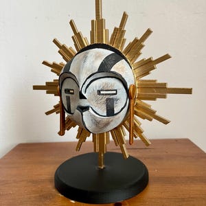 Può includere: Scultura di maschera solare decorativa con volto bianco, dettagli neri e dorati e design a raggi di sole dorati. La maschera ha una base nera ed è esposta su una superficie di legno. La maschera è alta circa 20 cm.