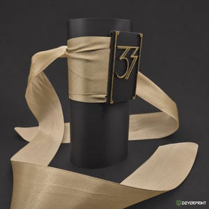 May include: A black cylindrical cuff featuring a wide, beige satin ribbon wrapped around the center. A rectangular black plaque with gold-colored trim is attached to the front, displaying the stylized gold-colored number 33 in a serif font.