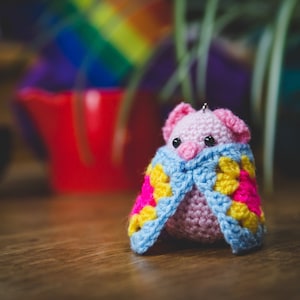 May include: A handmade, pink crocheted moth with black eyes and a blue and multicolored shawl. The shawl features yellow, pink, and blue squares. The moth is sitting on a wooden surface.