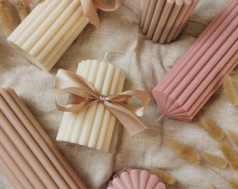 May include: An assortment of decorative candles in various shapes and colors. The candles are ribbed and feature shades of cream, beige, and pink. Some are tied with satin ribbons. The arrangement includes cylindrical, rectangular, and flower-shaped candles.