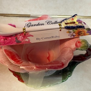 May include: A ceramic teacup and saucer set shaped like a pink flower with green leaves, accompanied by a matching spoon featuring a butterfly handle. The set is attached to a cardboard display card labeled Garden Collection By CornerRuby.