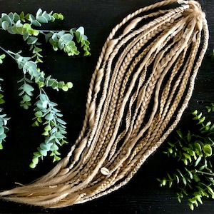 Könnte beinhalten: Ein Set aus blonden und braunen synthetischen Haarverlängerungen mit weißen und goldenen Akzenten. Die Verlängerungen sind geflochten und mit einem weißen Band zusammengebunden.