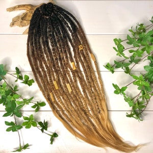 Könnte beinhalten: Ein Set langer, ombrefarbener brauner und blonder synthetischer Dreadlocks mit goldenen Perlen. Die Dreadlocks sind fächerförmig angeordnet und liegen auf einer weißen Holzoberfläche.