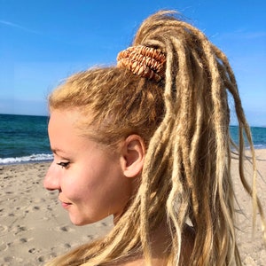 Könnte beinhalten: Eine Frau mit langen blonden Dreadlocks, die ein braunes Haargummi trägt. Das Haargummi ist aus natürlichen Materialien gefertigt und hat eine gewebte Textur.