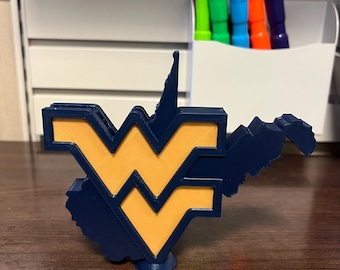 May include: A navy blue and gold 3D-printed sculpture of the West Virginia University logo, featuring the interlocking "WV" design. The sculpture is on a wooden surface, with a white container holding colorful markers in the background.