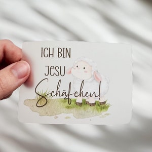Könnte beinhalten: Eine rechteckige Karte mit dem Text "ICH BIN JESU Schäfchen!" und einer Aquarellillustration eines Schafs. Das Schaf ist weiß mit rosa Ohren und Hufen und steht auf grünem Gras. Die Karte wird von einer Hand gehalten.