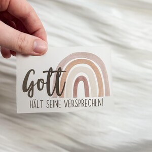 Könnte beinhalten: Ein Aufkleber mit dem deutschen Text "Gott hält seine Versprechen!" (Gott hält seine Versprechen) in dunkler brauner Schrift. Der Aufkleber zeigt einen Regenbogen in Braun- und Beigetönen.
