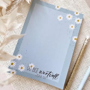 Könnte beinhalten: Hellblauer Notizblock mit Gänseblümchen-Blumendesign und dem Text "DU BIST wertvoll" und "JESAJA 43:3". Ein goldener Glitzerstift und ein hellblauer Bleistift liegen neben dem Notizblock. Der Notizblock hat einen hellblauen Rand.
