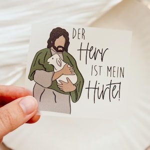 Könnte beinhalten: Ein Aufkleber mit einer Illustration einer Figur in einem grünen Gewand, die ein Lamm hält. Der Text lautet "DER Herr IST MEIN Hirte!" in schwarzer Schrift. Der Hintergrund ist eine weiche, neutrale Farbe.