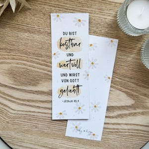 K&ouml;nnte beinhalten: Zwei wei&szlig;e Lesezeichen mit G&auml;nsebl&uuml;mchenmustern und deutschem Text. Der Text lautet "DU BIST kostbar UND wertvoll UND WIRST VON GOTT geliebt!" mit Bibelversangabe. Auf einem Holztablett.