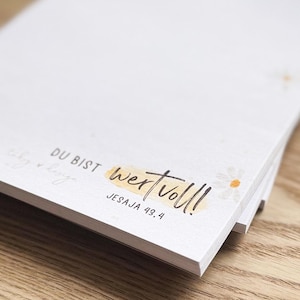 Könnte beinhalten: Ein Stapel weißer Notizblöcke mit dem Schriftzug "DU BIST wertvoll!" und der Referenz "JESAJA 43.4" in schwarzer Schrift. Das Wort "wertvoll" ist durch ein gelbes Rechteck hervorgehoben. Kleine Gänseblümchen-Illustrationen sind ebenfalls vorhanden.