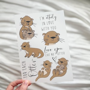 Könnte beinhalten: Eine weiße Karte mit Cartoon-Ottern in verschiedenen Posen. Der Text lautet "I'm otterly in love with you" und "Love you like no otter". Die Otter sind braun mit schwarzen Schnurrhaaren. Eine Herzgrafik ist ebenfalls enthalten.