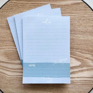 Könnte beinhalten: Ein Stapel von drei hellblauen Notizblöcken mit linierten Seiten. Das Wort "notes" ist in Kursivschrift oben geschrieben, und das Wort "wichtig" unten. Ein Papierflieger und ein Herz sind ebenfalls zu sehen.