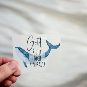 Könnte beinhalten: Ein Aufkleber mit einer blauen Aquarell-Wal-Illustration. Der Aufkleber hat den deutschen Text "Gott sieht dich überall!" in schwarzer Schrift. Der Wal hat einen weißen Bauch und eine blaue Schwanzflosse.