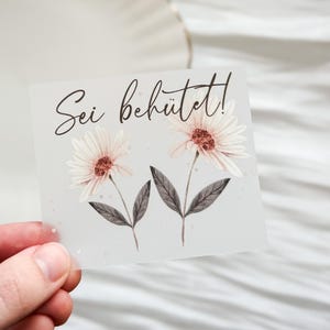 Könnte beinhalten: Eine quadratische, durchscheinende Karte mit zwei Aquarell-Gänseblümchen, weißen Blütenblättern und rotbraunen Zentren. Die Karte trägt den Text "Sei behütet!" in eleganter Schrift. Die Karte wird von einer Hand vor einem weichen, verschwommenen Hintergrund gehalten.