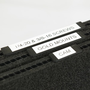 May include: Close-up of three black labels with white text on a black foam surface. The labels read "1/4-20 & 3/8-16 SCREWS", "GOLD MOUNTS", and "CAM".