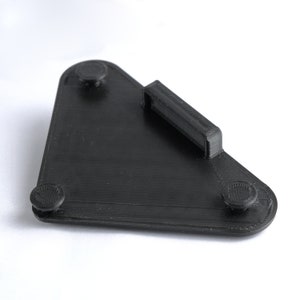 May include: A black, 3D-printed, triangular object with a handle and three circular feet. The object has a textured surface and is designed to hold a small item. The handle is rectangular and raised.