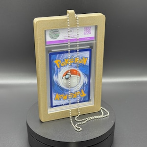 May include: A graded Pokémon trading card in a gold-coloured frame with a silver ball chain necklace. The card features a blue background with the Pokémon logo and a Poké Ball. The frame is rectangular and has a clear front.