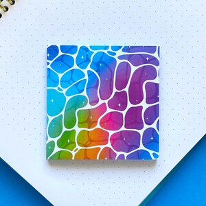Peut inclure: Un autocollant carré présentant un motif dégradé arc-en-ciel vibrant avec des contours organiques blancs en forme de cellules et de petites étoiles à quatre branches. L'autocollant est posé sur une page de carnet à points près d'une règle bleue.
