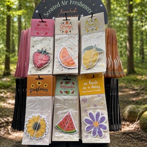 May include: A display of assorted scented air fresheners. The air fresheners are shaped like a strawberry, orange slice, lemon, sunflower, watermelon slice, and a daisy. The display includes the text "Scented Air Fresheners" and "Assorted Fragrances".