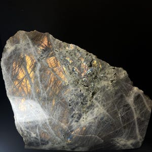 May include: A large, rough-cut labradorite gemstone with a dark grey base colour. The stone displays iridescent flashes of gold, blue, and green across its surface. The stone has a natural, unpolished appearance.