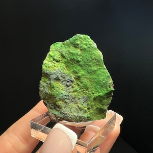May include: A green, irregularly shaped mineral specimen with a rough, textured surface. The specimen is displayed on a clear acrylic stand.