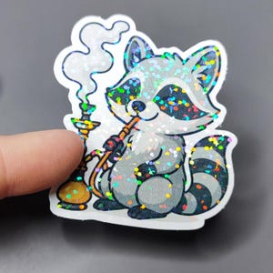 Könnte beinhalten: Ein glitzernder silberner und schwarzer Waschbär-Aufkleber mit einem bunten Glitzer-Finish. Der Waschbär hält eine Wasserpfeife und bläst Rauchringe.