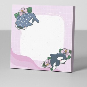 Könnte beinhalten: Ein quadratischer Notizblock mit hellrosa Gittermuster und weißer Mitte. Der Notizblock ist mit Cartoon-Illustrationen eines Walhais und eines Orcas verziert, die jeweils von Blumen und Blättern umgeben sind.