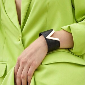 Puede incluir: Un brazalete de cuero negro con un acento en forma de V plateado. El brazalete se lleva en la muñeca.