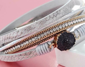 May include: A multi-layered bracelet featuring a grey leather strap, a gold chain, a row of clear rhinestones, and a grey cord. The bracelet has a black stone charm.