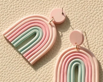 Könnte beinhalten: Ein Paar Regenbogen-Ohrringe zum Hängen. Die Ohrringe sind aus rosa, hellblauem und weißem Ton gefertigt und haben die Form von Regenbögen. Die Ohrringe haben ein schlichtes, minimalistisches Design.