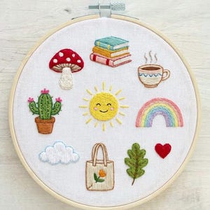 May include: A circular wooden embroidery hoop containing white fabric with hand-stitched designs including a red mushroom, a stack of three books, a steaming cup, a cactus, a smiling yellow sun, a rainbow, a cloud, a tote bag, a leaf, and a heart.