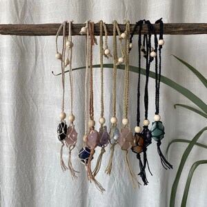 May include: A collection of eight crystal necklaces with macrame knotting and wooden beads. The crystals are various colors, including green, purple, pink, and brown. The necklaces are hanging from a branch.