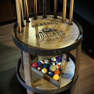 May include: A wooden billiard club stand with the text "Brannon Billiard Club" on the top. The stand has a circular design with metal bands and holds pool cues and a set of billiard balls in a wooden triangle.