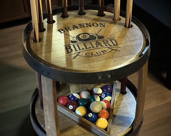 May include: A circular wooden billiard cue rack with a dark metal rim. The top surface features the engraved text BRANNON BILLIARD CLUB and an 8-ball graphic. The lower shelf holds a triangular wooden rack filled with colorful numbered billiard balls.