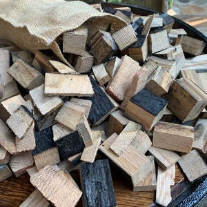 May include: A burlap sack spills out a variety of wood chips, some light brown and some dark brown with black charring, onto a wooden surface. The chips are likely for smoking food.
