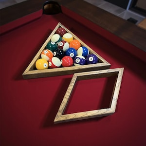 May include: A wooden triangle rack holds a set of numbered billiard balls, arranged on a red felt pool table. A wooden diamond-shaped rack sits below the triangle. The balls are various colors, including red, yellow, and blue.