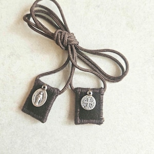 May include: Two small, dark brown scapulars with silver pendants, joined by a brown cord. One pendant depicts a religious figure, the other a cross. The scapulars are made of dark brown fabric.