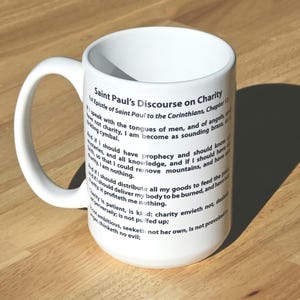 May include: White ceramic mug with a curved handle. The mug features the text "Saint Paul's Discourse on Charity" and excerpts from the 1st Epistle to the Corinthians, Chapter 13, in black lettering. The mug is on a wooden surface.
