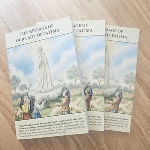 May include: Three copies of a book titled "The Message of Our Lady of Fatima." The cover features an illustration of a religious figure with children and sheep. The books are stacked on a wooden surface.