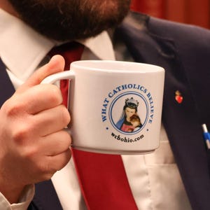 May include: White ceramic mug with a blue and white logo that says "What Catholics Believe" and "wcbohio.com". The logo features a depiction of the Virgin Mary holding a baby Jesus.