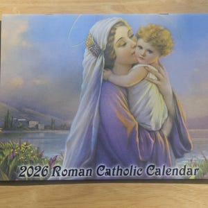 以下が含まれることがあります： 2026年のローマカトリックカレンダー。白いベールをかぶった女性が子供にキスをしている絵が描かれています。女性は紫色のローブを着ており、子供は巻き毛のブロンドの髪をしています。背景には湖と建物が描かれています。