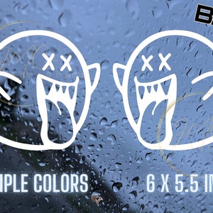 May include: Two white vinyl decals of a ghost with a mischievous grin and crossed-out eyes. The decals are 6 inches by 5.5 inches and are available in multiple colors.