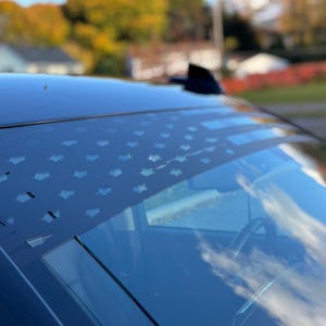 May include: A black and white American flag decal on the windshield of a car. The flag is displayed horizontally across the top of the windshield.