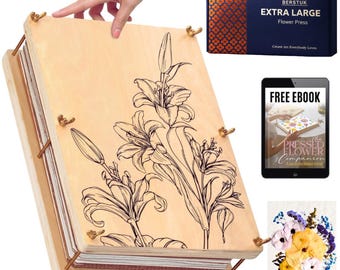 May include: A light-colored wooden flower press featuring a black line drawing of lilies on the top cover. The press is secured with four metal bolts and wing nuts. Included are a dark blue box labeled BERSTUK EXTRA LARGE Flower Press, a tablet displaying a free ebook, and a small card showing a colorful arrangement of pressed flowers.