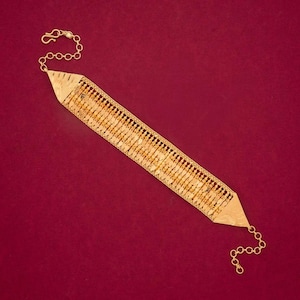 Könnte beinhalten: Ein goldfarbenes Armband mit geometrischem Design. Das Armband hat rechteckige Glieder, die durch kleinere dekorative Elemente verbunden sind. An den Enden sind eine Kette und ein Verschluss angebracht. Der Hintergrund ist ein tiefes Rot.