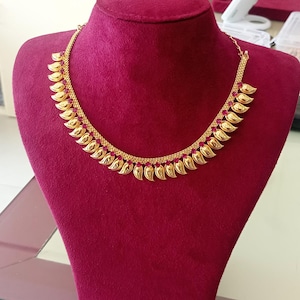 May include: A gold-colored necklace with a leaf-like design, displayed on a red velvet bust. The necklace features a delicate chain and multiple curved, textured leaf pendants. The background is slightly blurred, highlighting the jewelry.