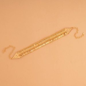 Può includere: Una collana girocollo dorata con un design geometrico. La collana presenta una serie di piccoli pezzi rettangolari dorati collegati in fila. Ha una chiusura a catena e chiusura. La lunghezza è di circa 30 cm.